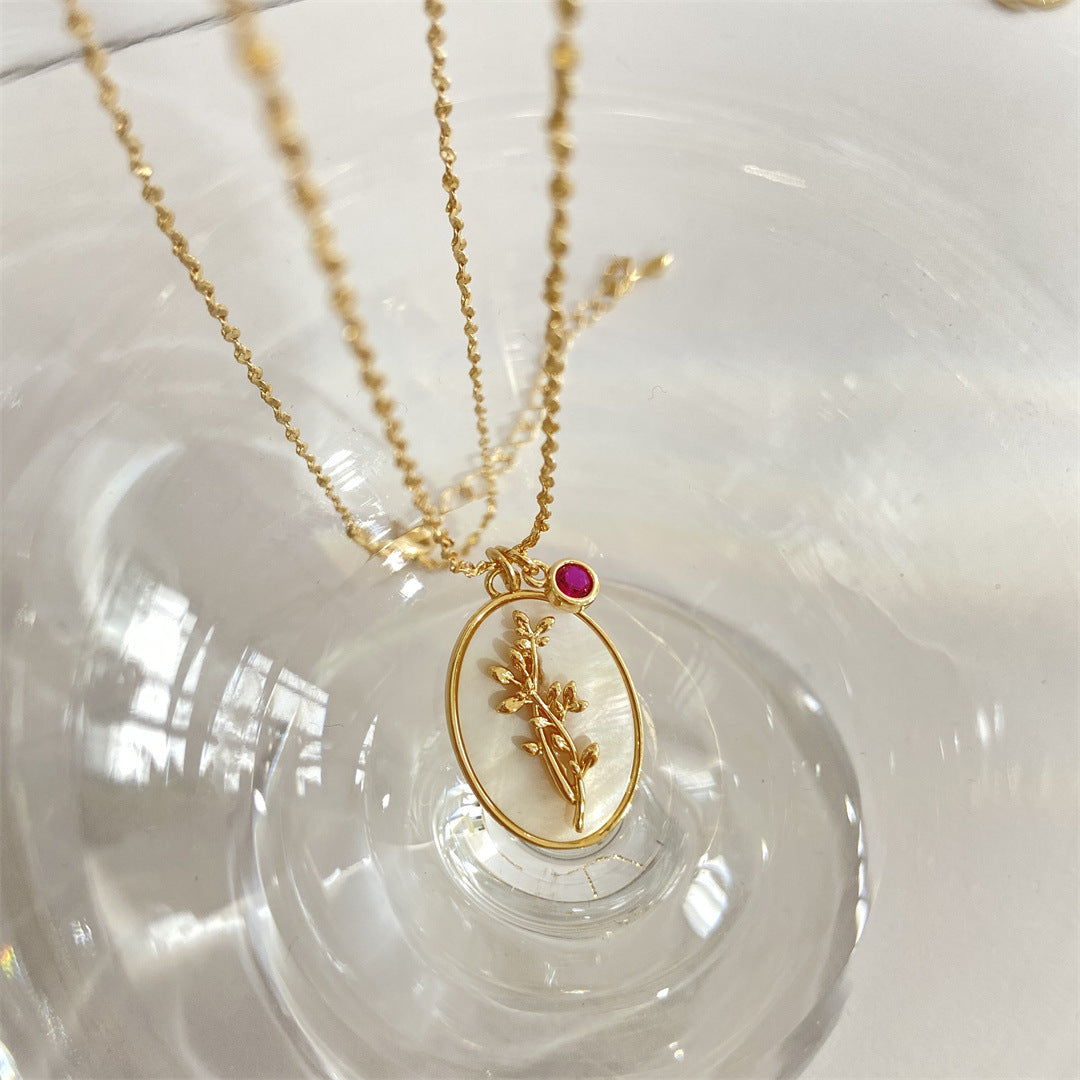Pendentif ovale en nacre avec une branche dorée et une petite pierre rouge, monté sur une chaîne fine en or.