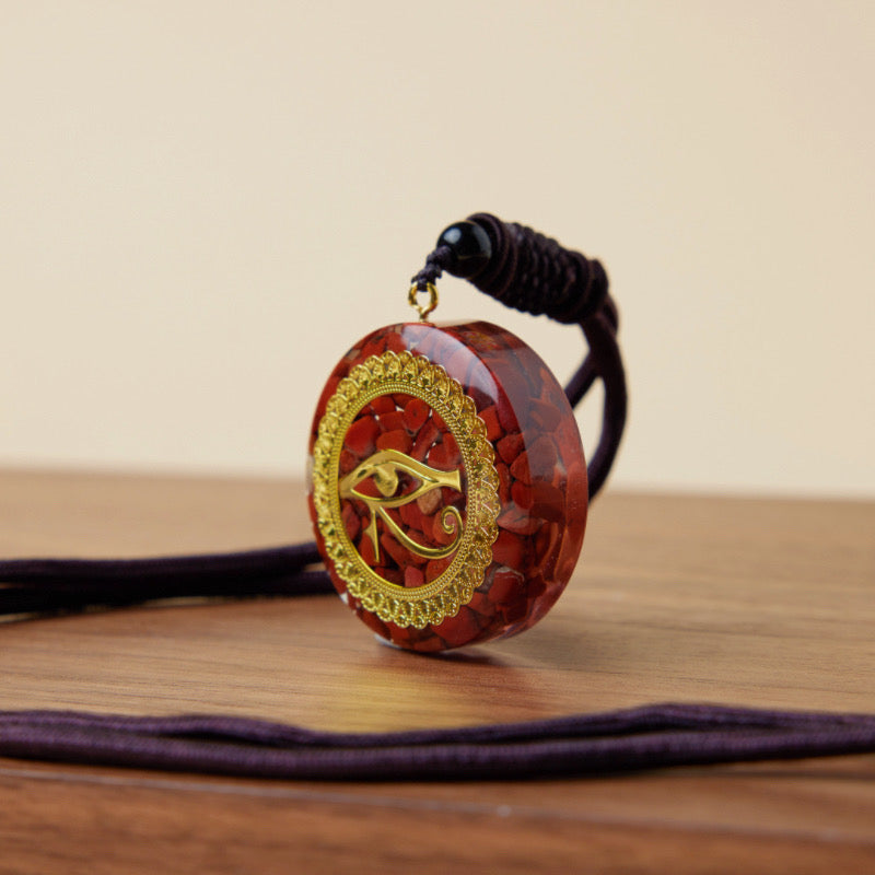 Pendentif rond en orgonite rouge incrusté de petits éclats de jaspe, orné d'un symbole doré de l'œil d'Horus au centre, suspendu à un cordon violet.