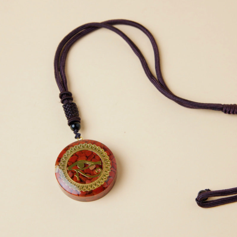 Pendentif rond en orgonite rouge incrusté de jaspe et orné du symbole de l'œil d'Horus, suspendu à un cordon violet.
