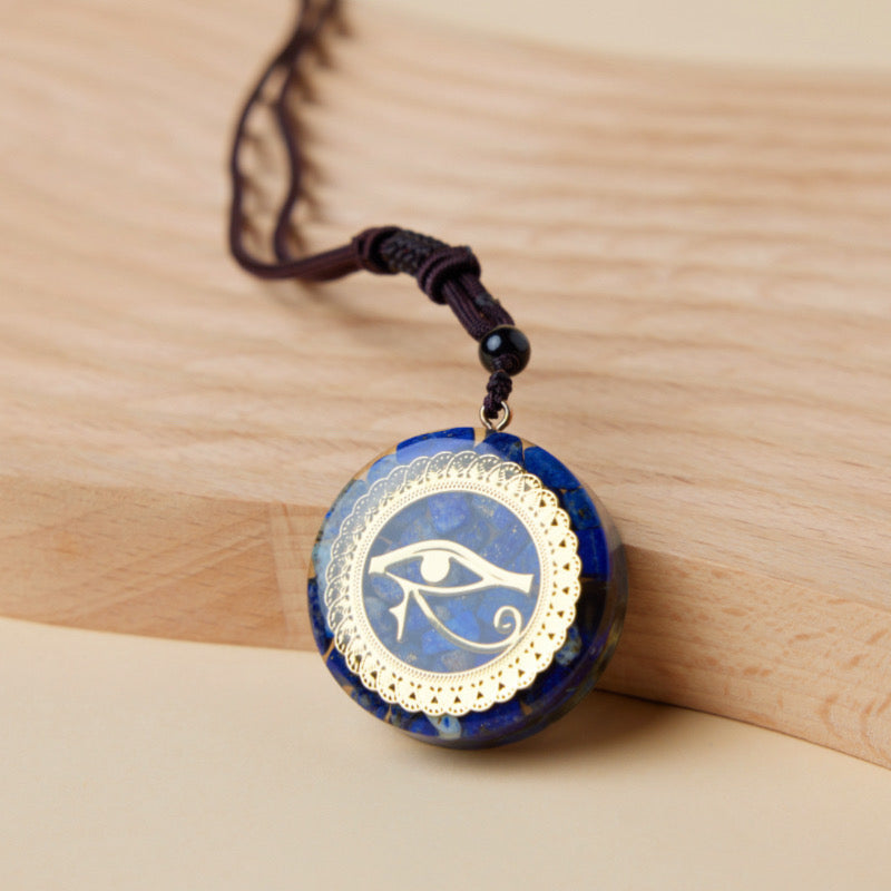 Pendentif Orgonite rond bleu profond avec un œil d'Horus doré au centre, bordé d'une dentelle dorée, sur un fond en bois clair.