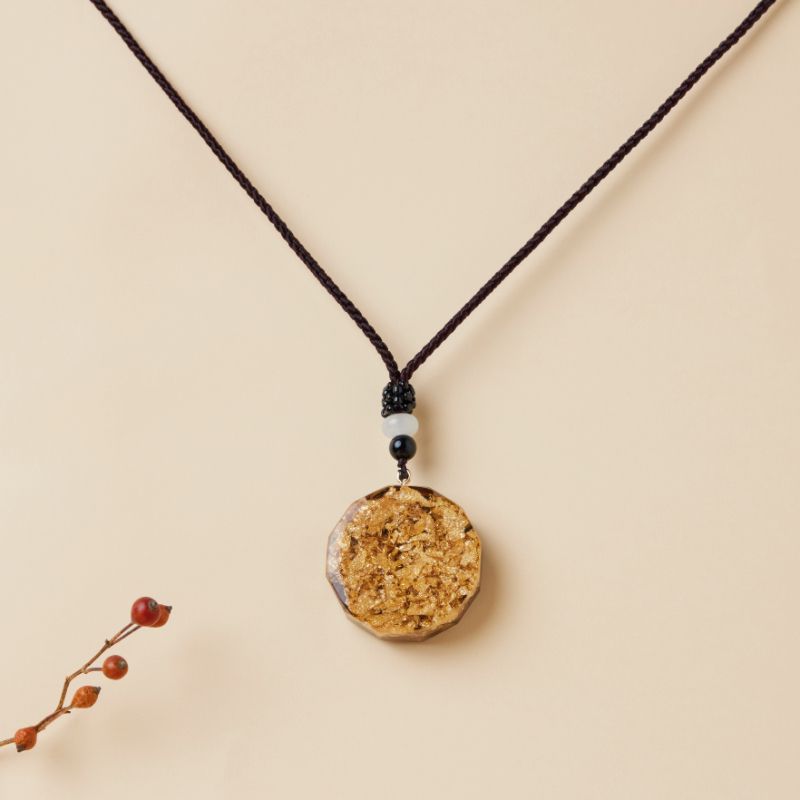 Pendentif rond en orgonite avec incrustations dorées, monté sur un cordon marron, orné de perles noires et blanches.
