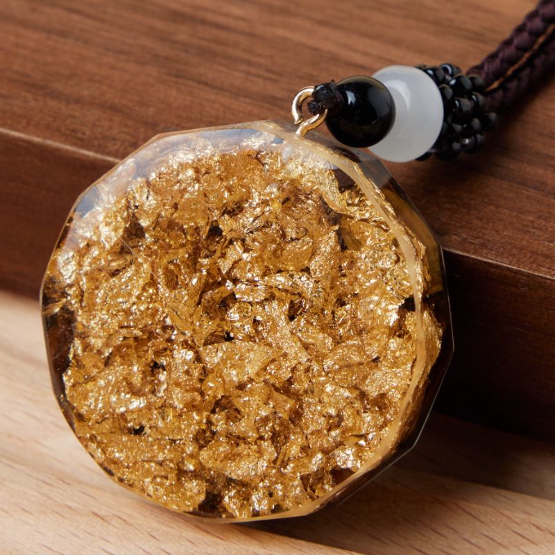 Pendentif orgonite octogonal incrusté de feuilles d'or, avec une cordelette marron et des perles noires et blanches.