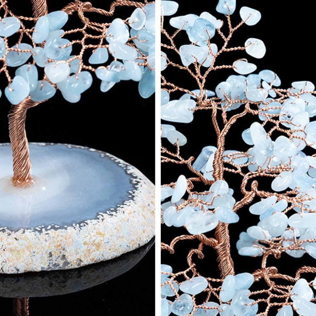 Arbre de vie "Sérénité" artisanal avec des feuilles en aigue-marine naturelle et un tronc en fil de cuivre, reposant sur une base en agate bleu clair veiné.