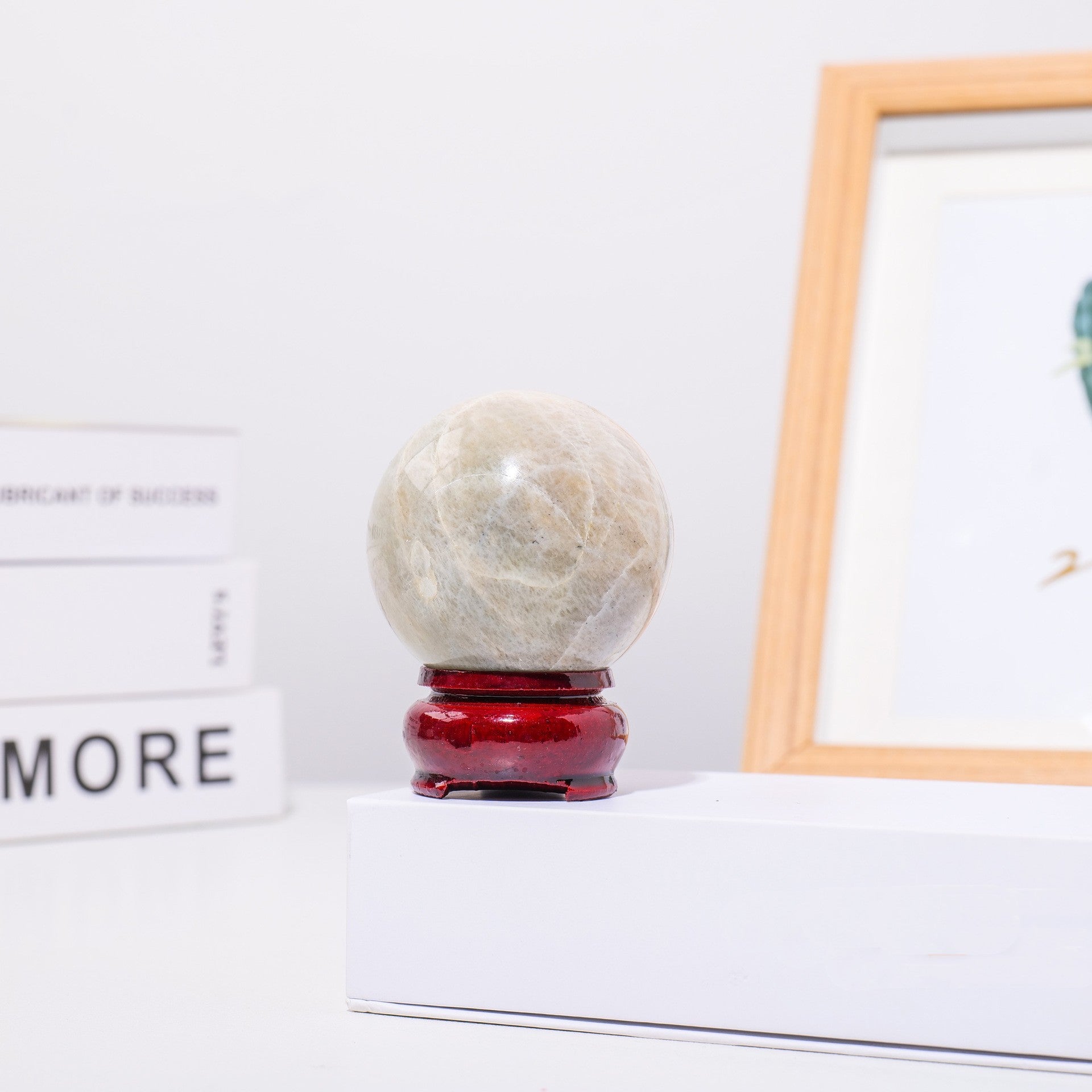 Une sphère en pierre de lune naturelle veinée de blanc et de gris, posée sur un socle rouge verni, avec un cadre photo en bois en arrière-plan.