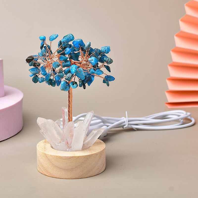 Arbre de vie lumineux avec des pierres bleues et une base en cristal de quartz sur un socle en bois, relié à un cùble d'alimentation blanc.