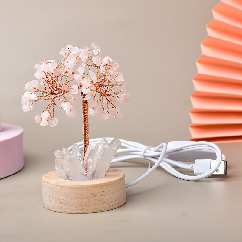 Arbre de vie lumineux avec des feuilles en quartz rose et une base en bois, orné de cristaux de roche et doté d'un cùble USB blanc.