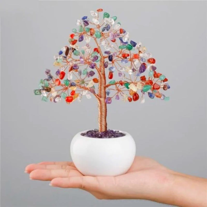 Arbre de vie décoratif aux pierres multicolores des 7 chakras, posé dans un pot blanc tenu dans une main.