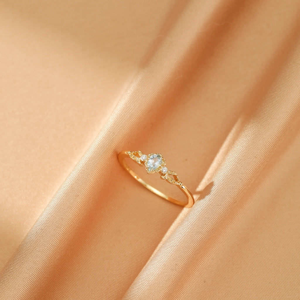Bague en argent plaqué or 14K avec une aigue-marine centrale et de petits cristaux scintillants sur le côté, posée sur un tissu satiné couleur pêche.
