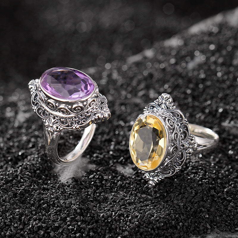 Deux bagues artisanales en argent massif ornées de pierres précieuses ovales, l'une avec une améthyste violette et l'autre avec une citrine jaune, sur un fond sombre.