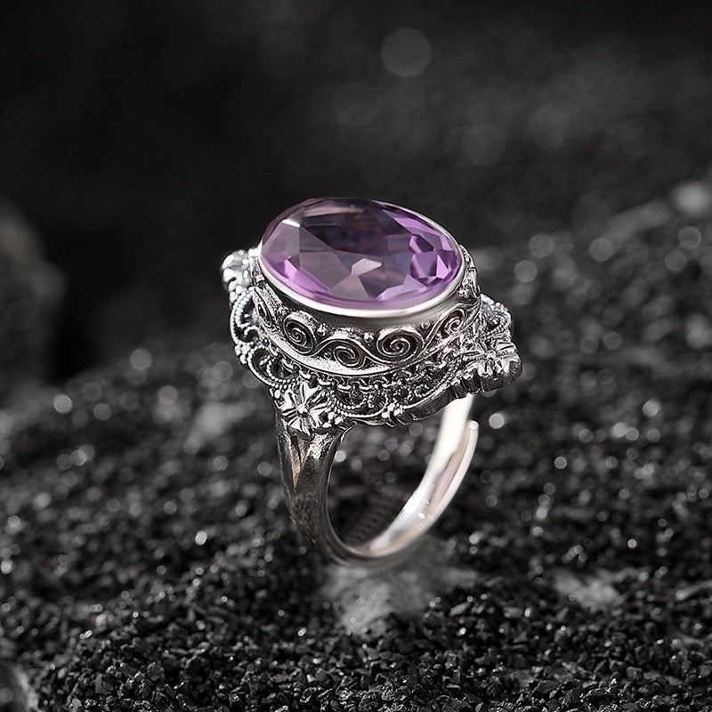 Bague Princesse Mystérieuse sertie d'une grosse améthyste ovale taillée, surmontée d'une monture complexe en argent vieilli aux motifs floraux et volutes.