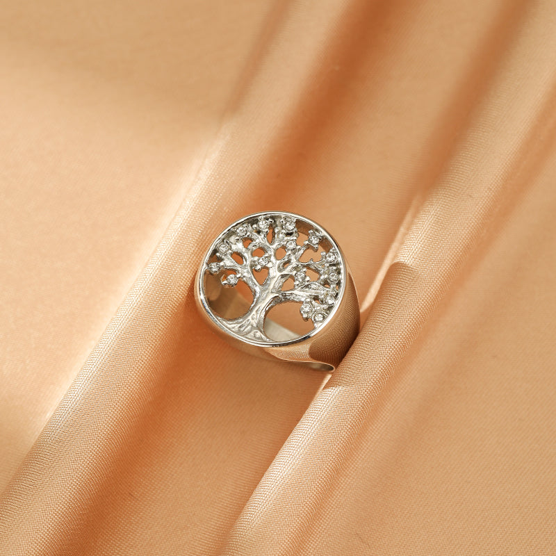 Bague argentée avec motif Arbre de Vie ajouré et incrusté de petites pierres scintillantes, sur un fond satiné couleur pêche.