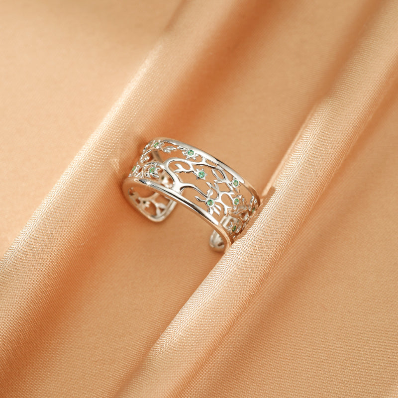 Bague ouverte en argent 925 avec motif d'arbre de vie ajouré et petites pierres vertes incrustées, sur fond de tissu satiné couleur pêche.