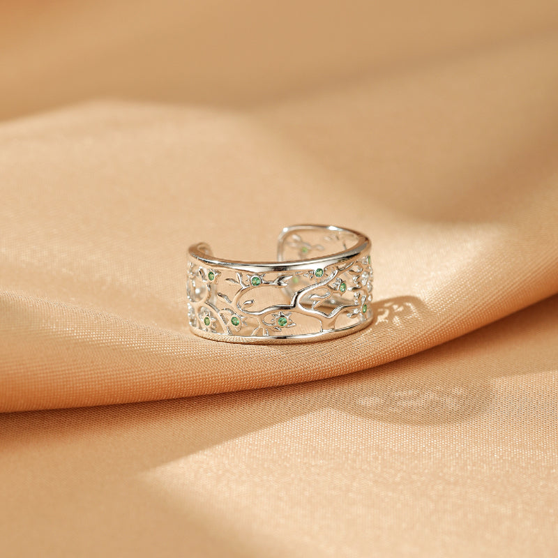 Bague ouverte en argent 925 ornée d'un motif d'arbre de vie ajouré et de petites pierres vertes, posée sur un tissu satiné beige.