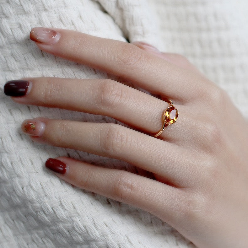 Une bague en argent plaqué or avec une citrine ovale orange est portée à l'annulaire d'une main aux ongles bordeaux et nude pailletés.