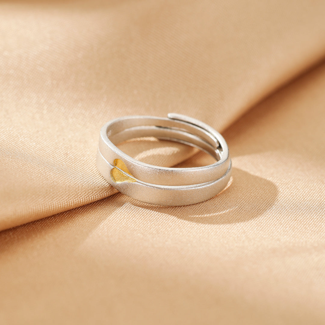 Bague de couple en argent avec un cœur doré entrelacé, sur fond de tissu satiné.