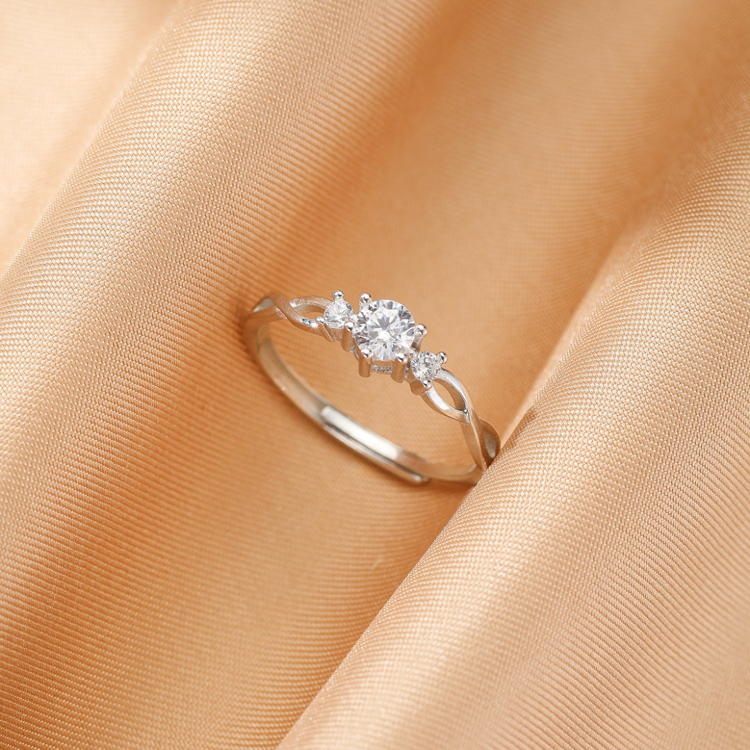 Bague de fiançailles en argent avec trois pierres scintillantes, dont une centrale plus grande, sur un fond de tissu satiné doré.