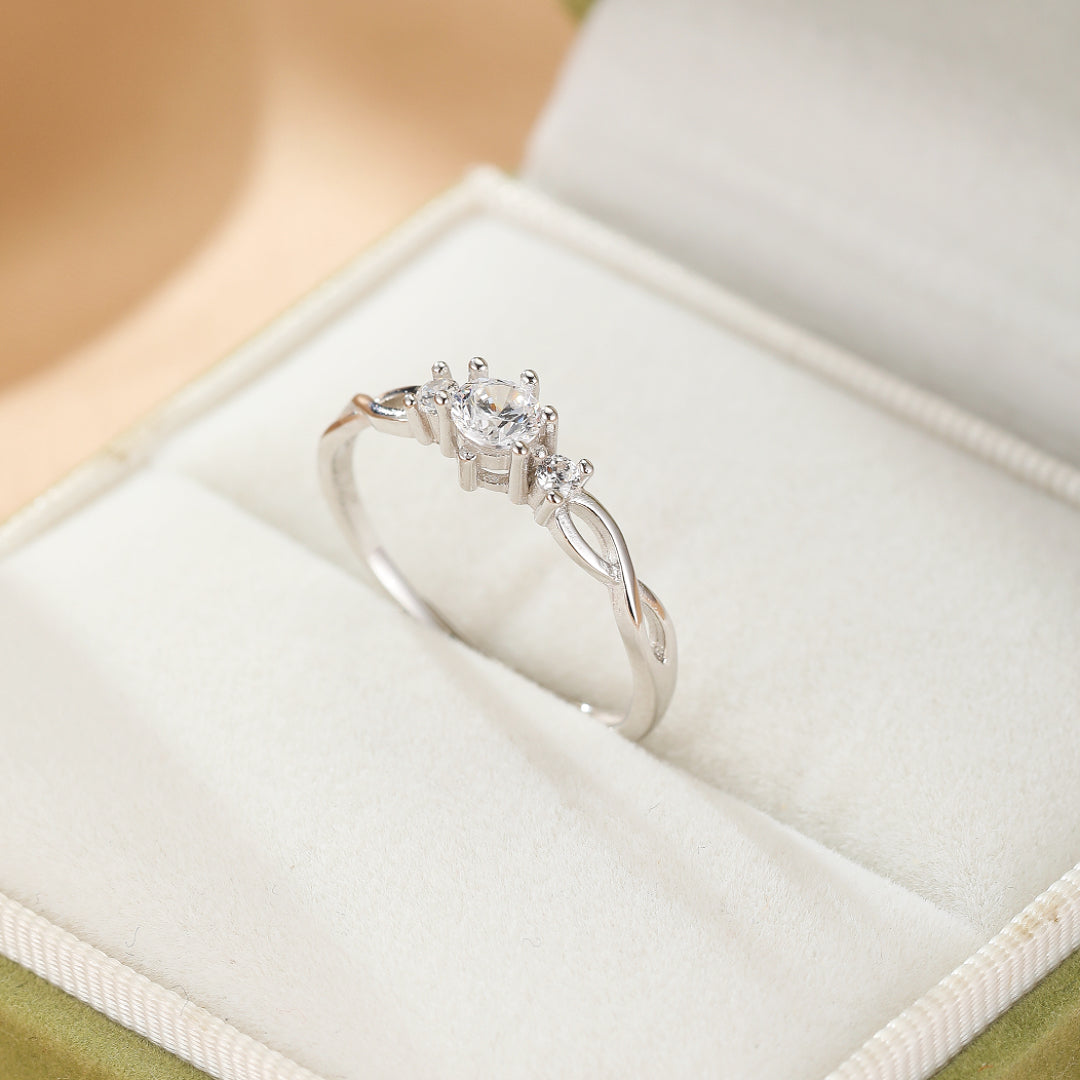 Bague fine en argent avec un design torsadé et une pierre centrale entourée de deux petites pierres scintillantes, présentée dans un écrin.