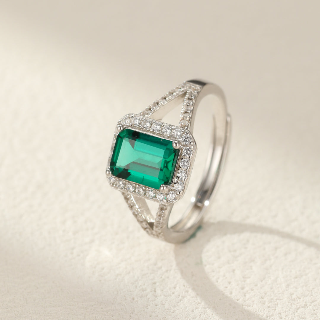 Bague en argent 925 avec une émeraude rectangulaire centrale entourée de petits cristaux brillants et une monture ajourée.