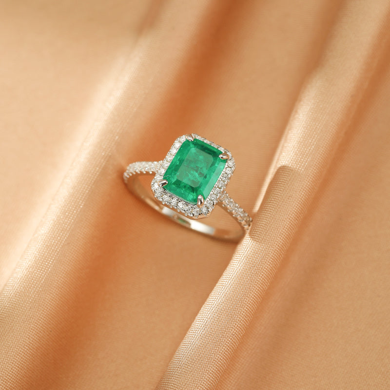 Bague en argent sertie d'une émeraude rectangulaire entourée de petits diamants, sur fond satiné couleur pêche.