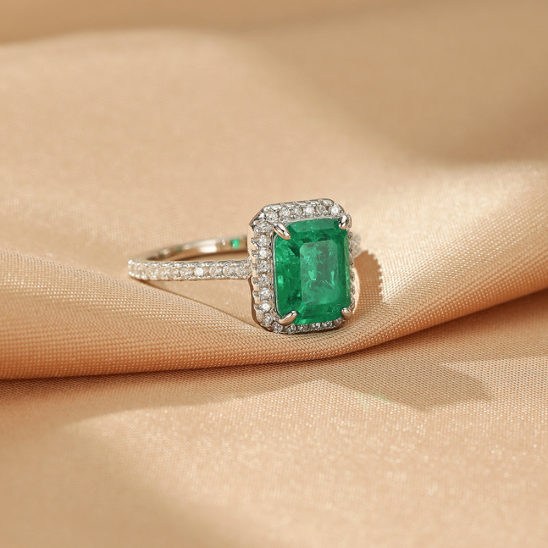 Bague en argent sertie d'une émeraude centrale taille émeraude, entourée d'un halo d'éclats de diamants, posée sur un tissu satiné couleur champagne.
