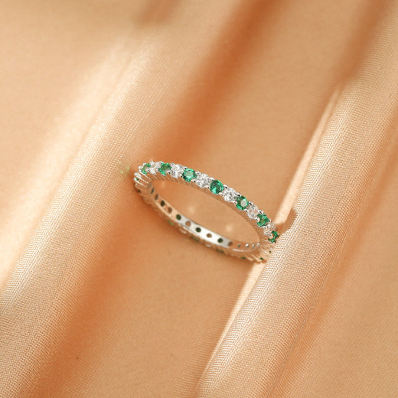 Bague alliance en argent sertie de petites pierres rondes vertes et blanches alternées, posée sur un tissu satiné beige.
