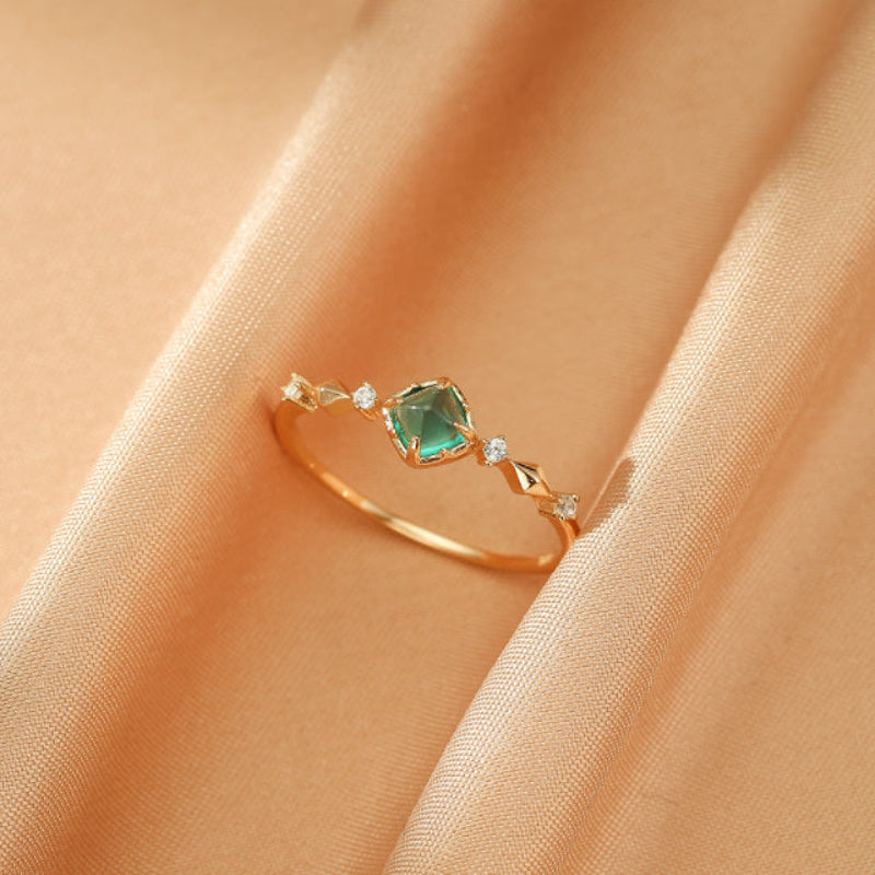 Bague Émeraude – Éclat Précieux, Argent Plaqué Or 14K. Une bague délicate en argent plaqué or 14K avec une émeraude carrée centrale et des accents de petits diamants. La bague repose sur un fond de tissu satiné beige.