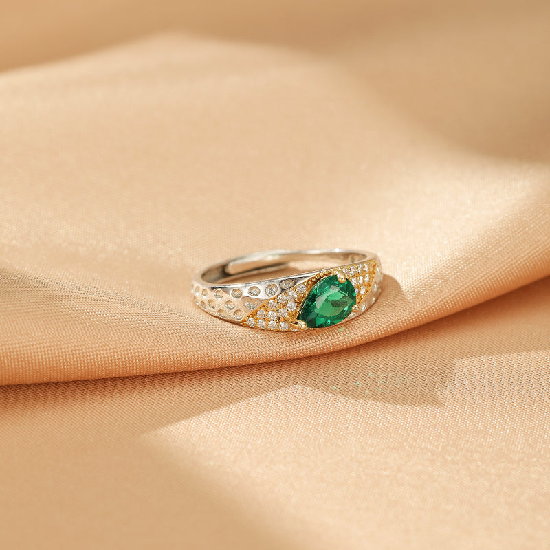 Bague émeraude en argent 925 "Larme d'Émeraude" avec une pierre verte en forme de larme sertie de petits cristaux brillants sur un fond satiné couleur pêche.