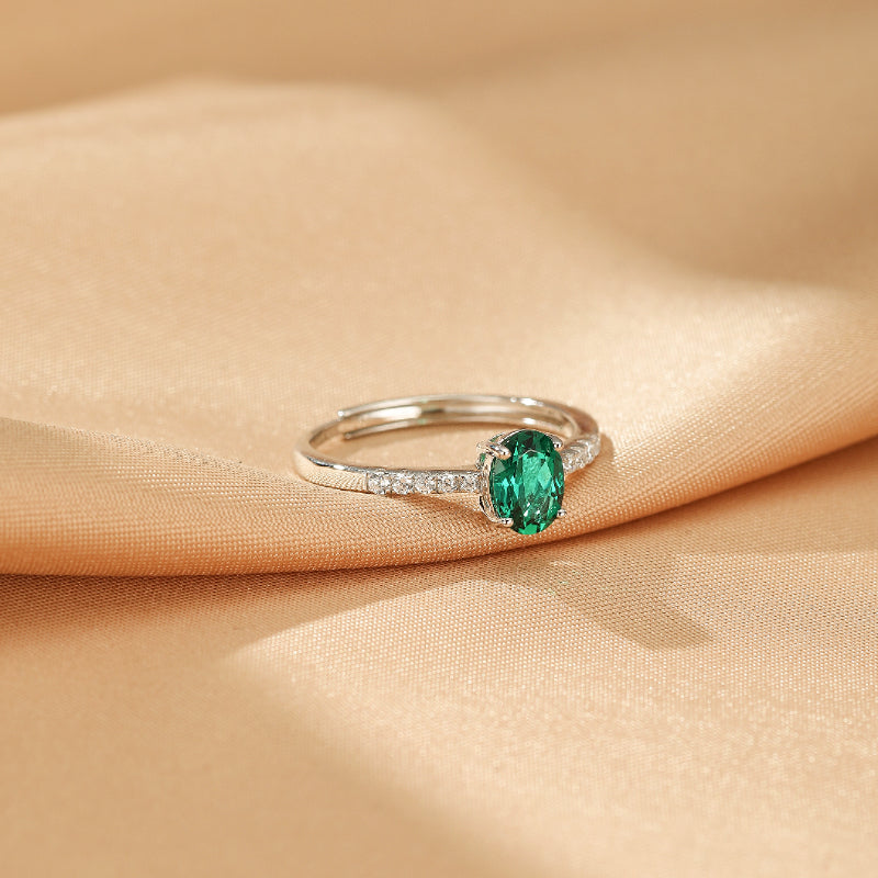 Bague ajustable en argent 925 avec une émeraude ovale verte centrale et un pavage de petits diamants sur l'anneau, posée sur un tissu satiné beige.