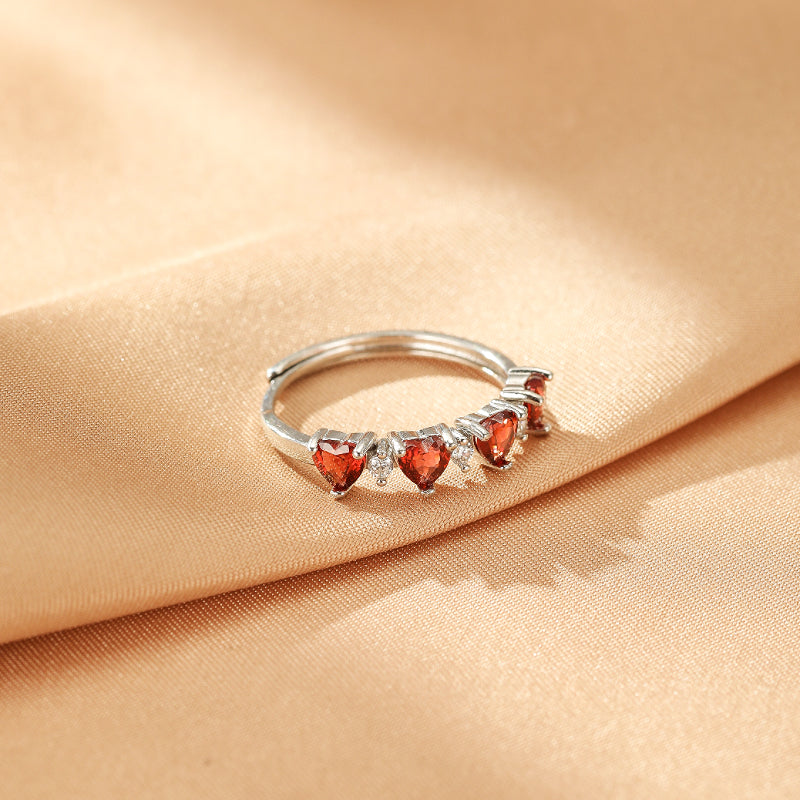 Bague ajustable en argent ornée de plusieurs cœurs de grenat rouge et de petites pierres d'imitation blanches, posée sur un tissu satiné couleur champagne.
