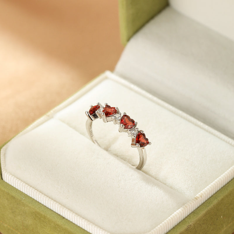 Bague en argent ornée de cinq cœurs de grenat rouge et de petites pierres blanches, présentée dans une boîte à bijoux verte.
