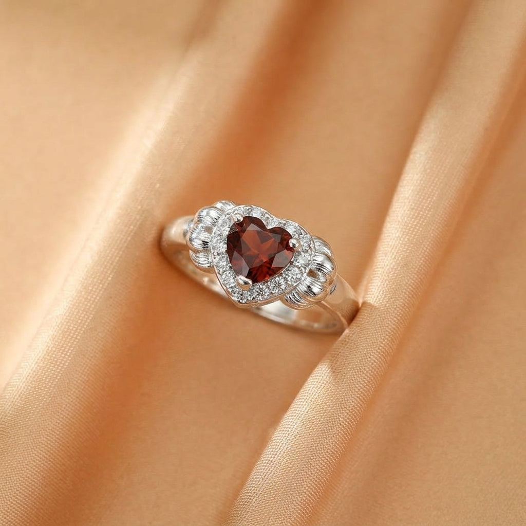 Bague en argent avec un grenat en forme de cœur entouré de petites pierres scintillantes, posée sur un tissu satiné doré.