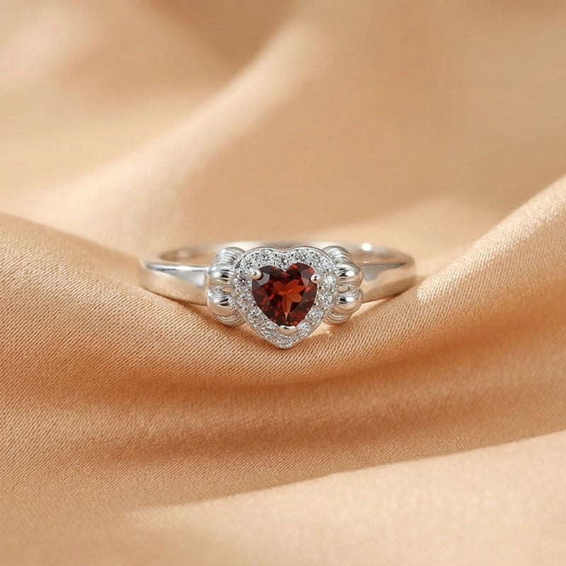 Bague en argent avec un grenat en forme de cœur entouré de petites pierres blanches scintillantes, posée sur un tissu satiné couleur pêche.