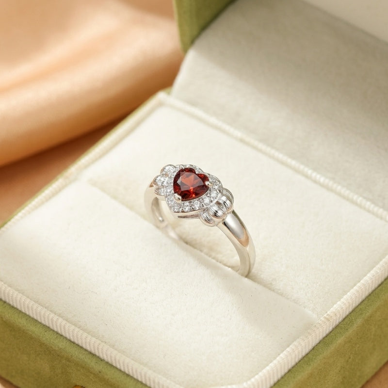 Bague en argent sterling avec un grenat en forme de cœur entouré de petites pierres blanches scintillantes, présentée dans une boîte à bijoux verte.