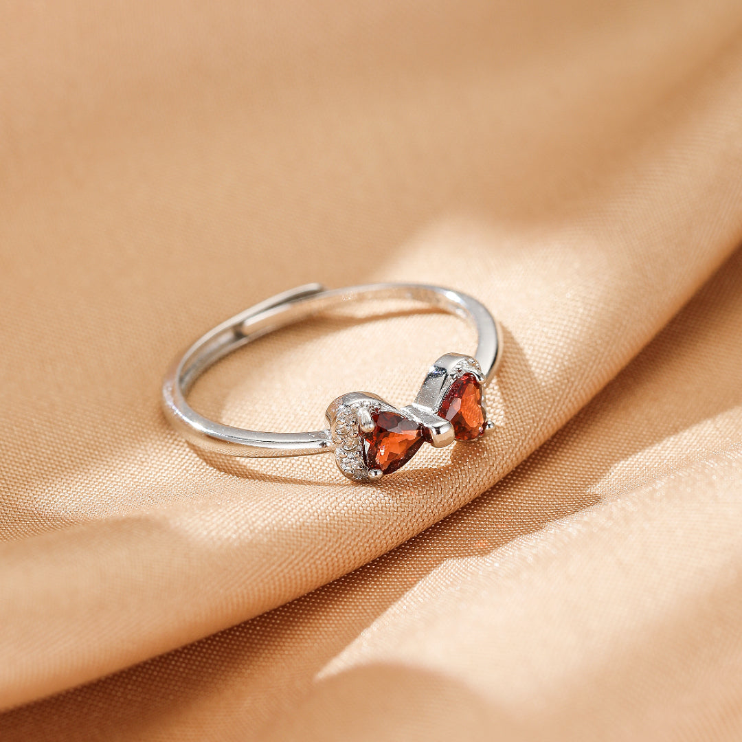 Bague en argent avec un nœud orné de grenats en forme de cœur et de petites pierres blanches scintillantes, posée sur un tissu satiné doré.