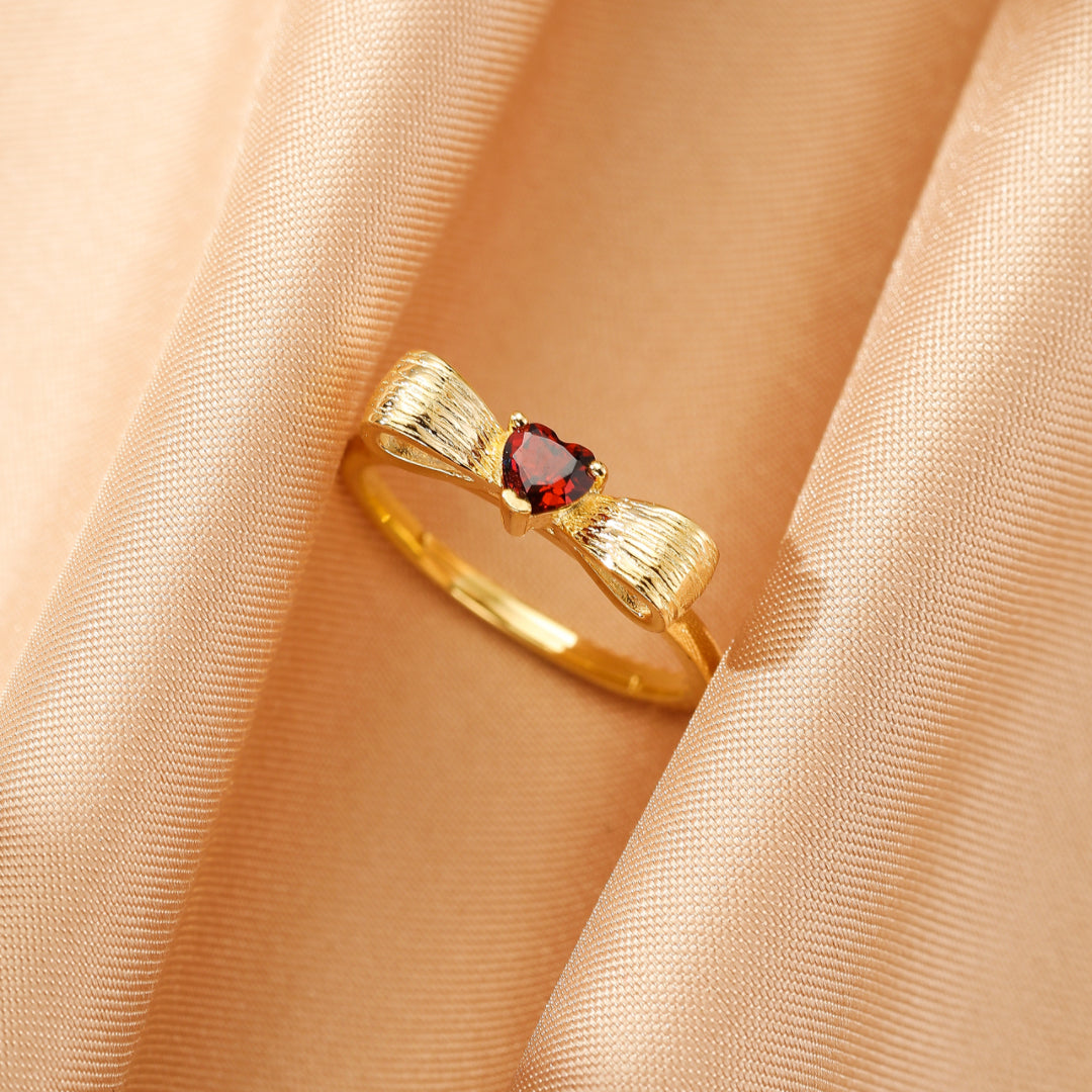 Bague nœud en argent plaqué or sertie d'un grenat en forme de cœur, sur un fond de tissu satiné couleur pêche.