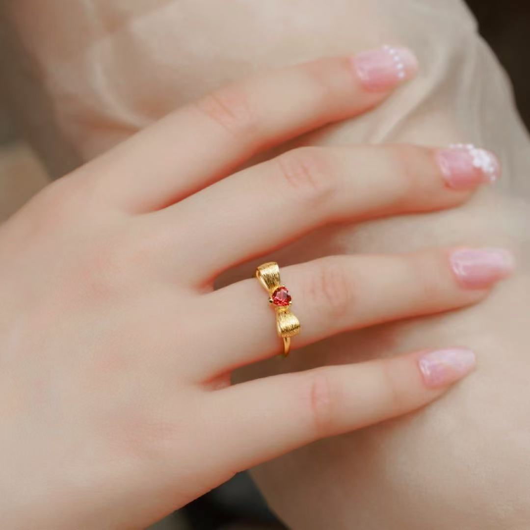 Bague nœud en argent plaqué or sertie d'une pierre grenat rouge en forme de cœur, portée sur l'annulaire d'une main aux ongles manucurés.