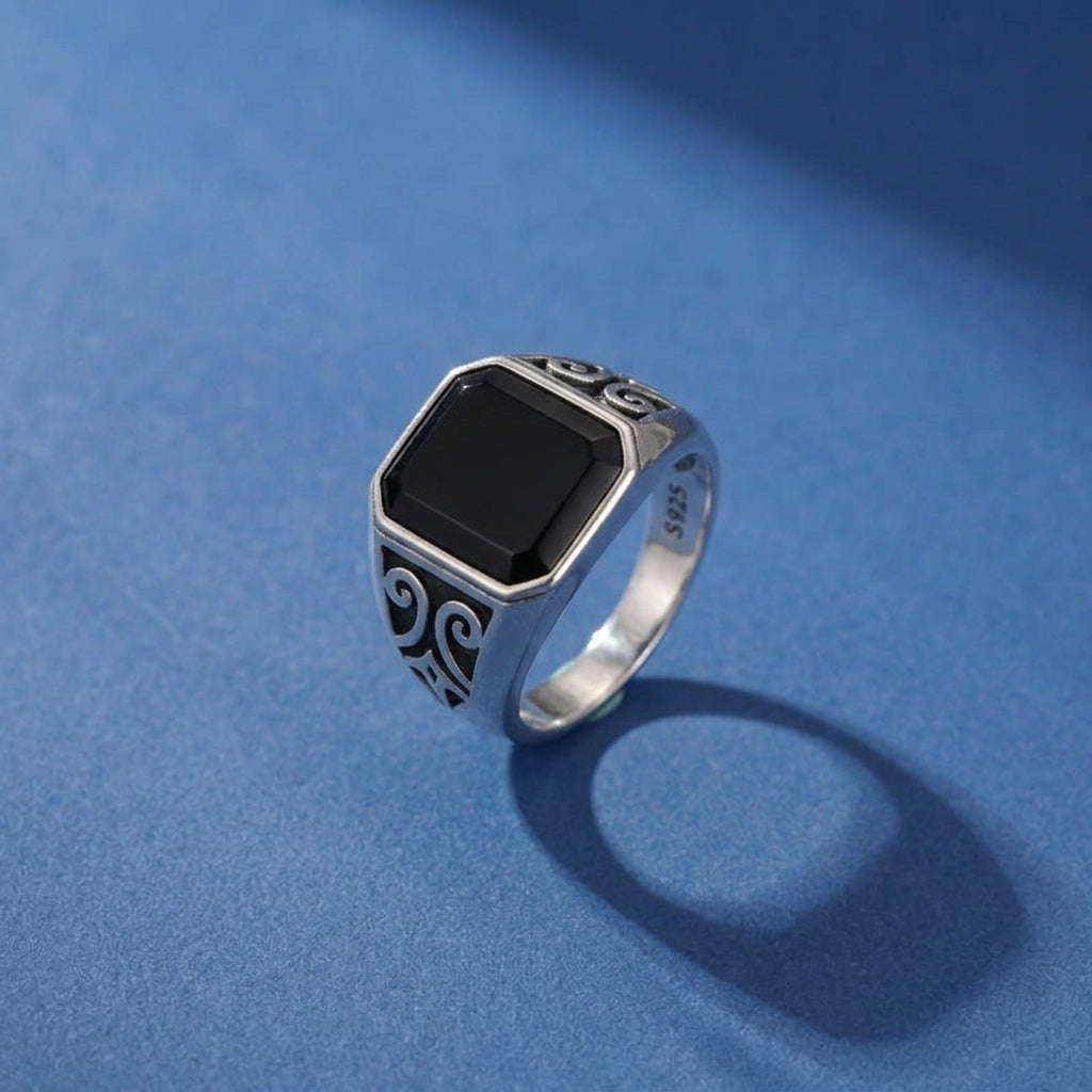 Bague pour homme en argent sterling 925 avec une pierre d'onyx noir rectangulaire et des détails sculptés sur les côtés, sur fond bleu.
