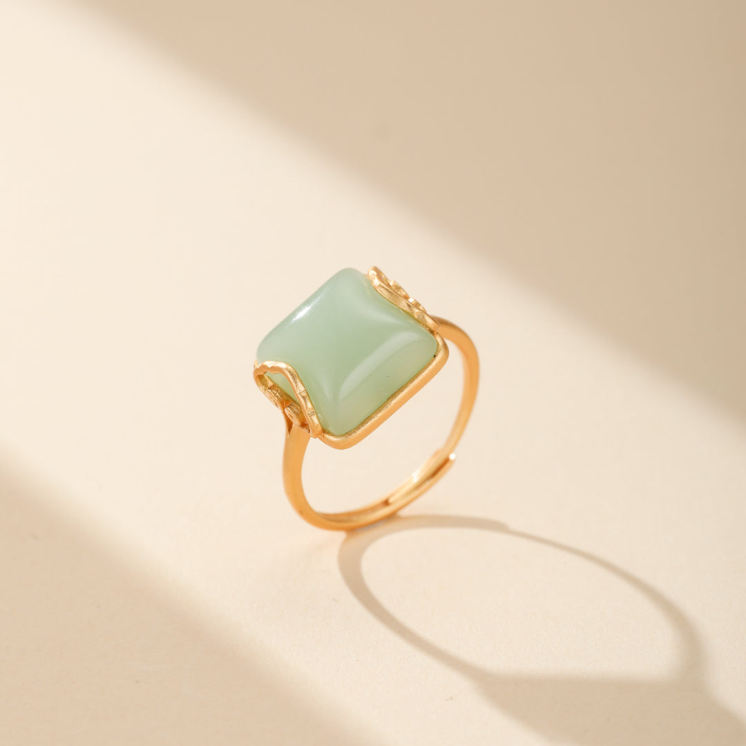 Gros plan sur la Bague Jade 'Essence d'Abondance' en plaqué or, mettant en valeur une pierre de jade vert foncé carrée flanquée de nuages dorés. Un bijou intemporel combinant tradition et modernité, évoquant des forêts mystiques.