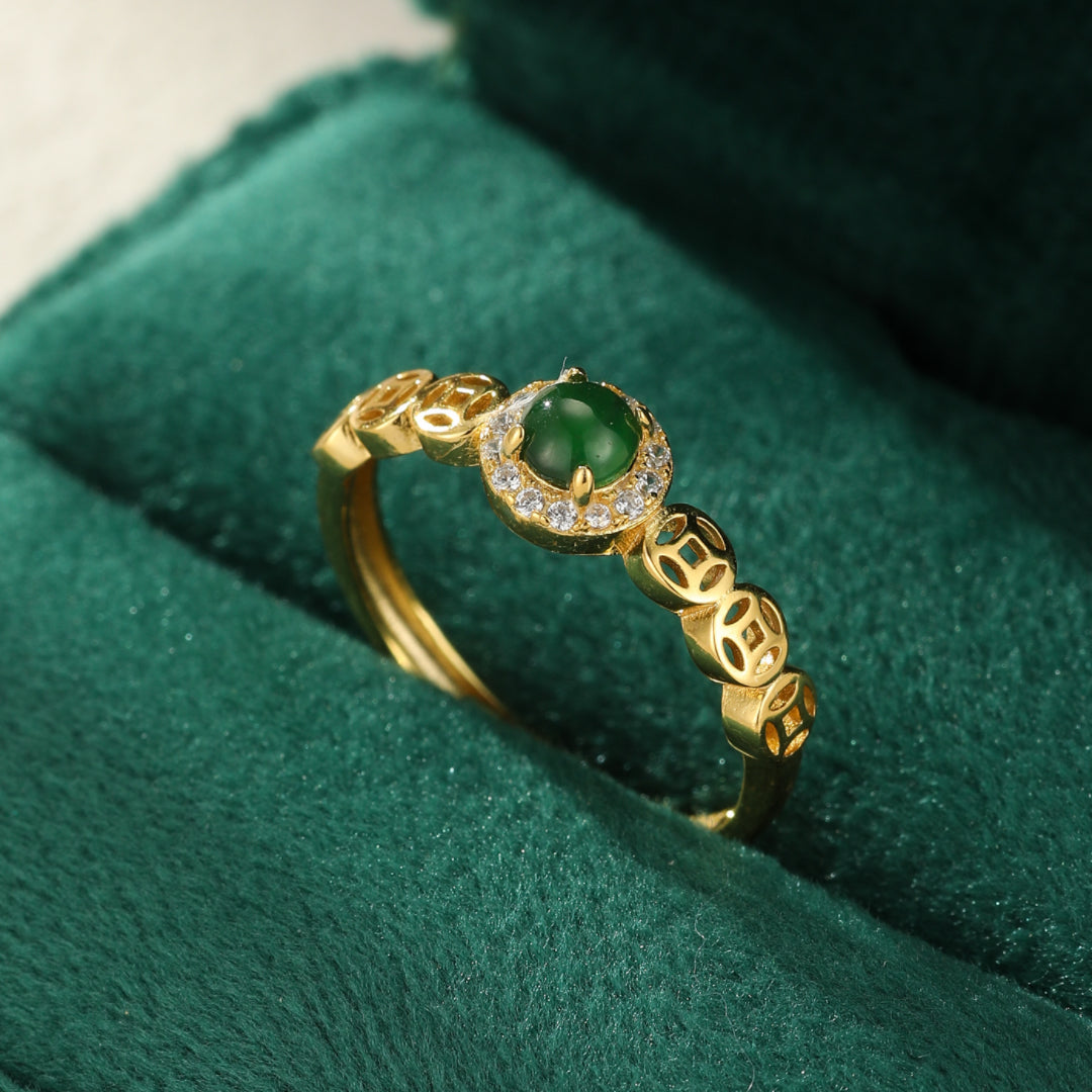 Bague en argent plaqué or sertie d'une pierre de jade verte ronde entourée de petits cristaux, avec une chaîne ornée de motifs de pièces chinoises ajourées, posée sur un fond vert en velours.