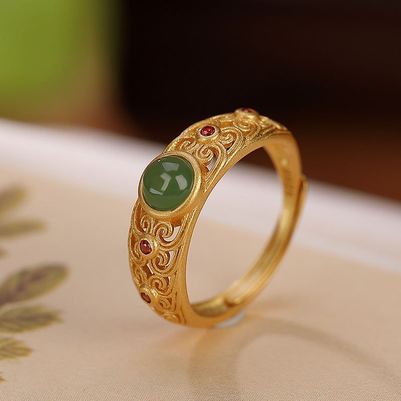 Bague en argent plaqué or sertie d'un cabochon de jade vert et de petites pierres rouges, avec des motifs gravés complexes.