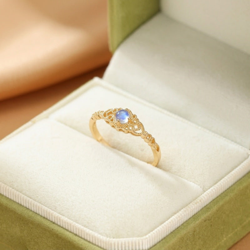 Bague en or avec une pierre de lune bleue centrale et des détails ouvragés sur le côté, présentée dans une boîte à bijoux verte.
