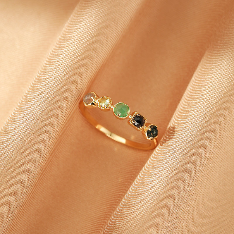 Bague en or sertie de cinq pierres rondes de couleurs variées : labradorite, chrysoprase et péridot, sur un fond de tissu satiné couleur pêche.