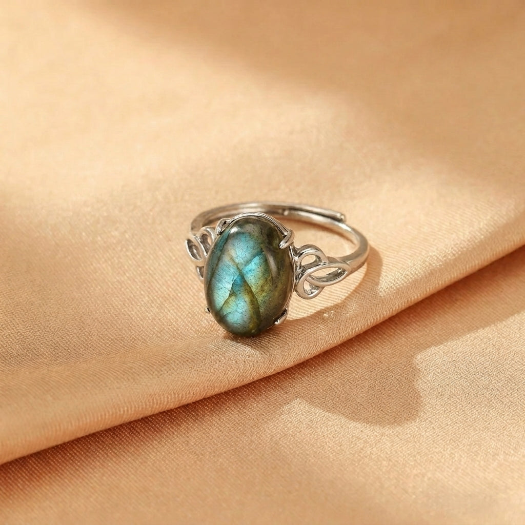 Bague ajustable en argent avec une pierre de labradorite ovale aux reflets bleus et verts, sur une monture ornée de volutes.