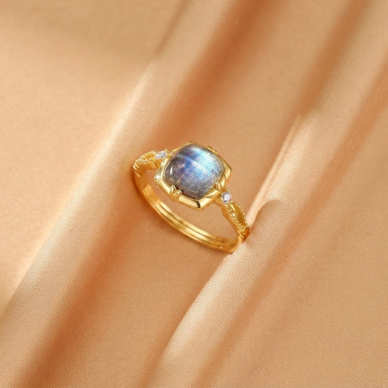 Bague en argent plaqué or avec une labradorite carrée aux reflets bleus et violets, ornée de petites pierres de zircon sur l'anneau torsadé.