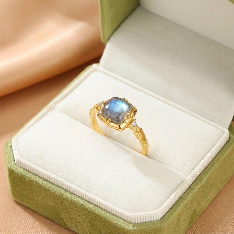 Bague Éclat Secret en argent plaqué or avec une labradorite carrée bleue et deux petites pierres blanches sur les côtés, présentée dans un écrin vert.