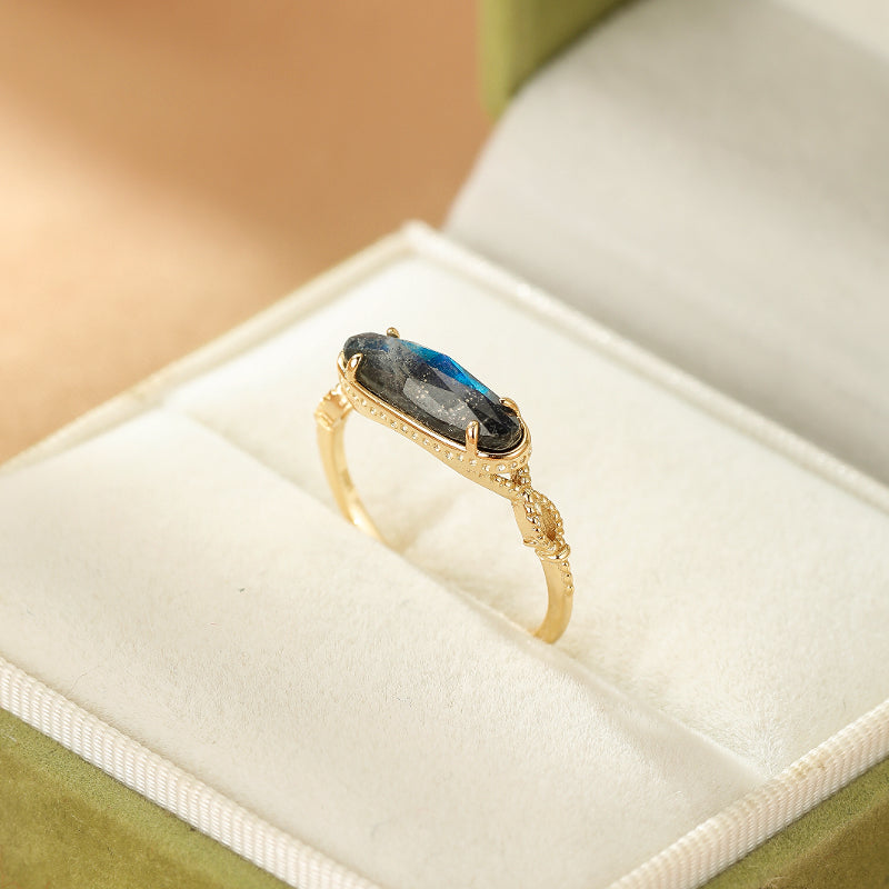 Bague en argent plaqué or avec une pierre de labradorite allongée aux reflets bleus et verts, montée sur un anneau fin orné de détails tressés.