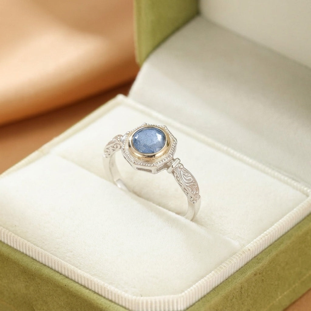 Bague en argent plaqué or sertie d'une pierre de labradorite bleue aux reflets profonds, ornée de motifs gravés sur l'anneau, présentée dans une boîte verte.