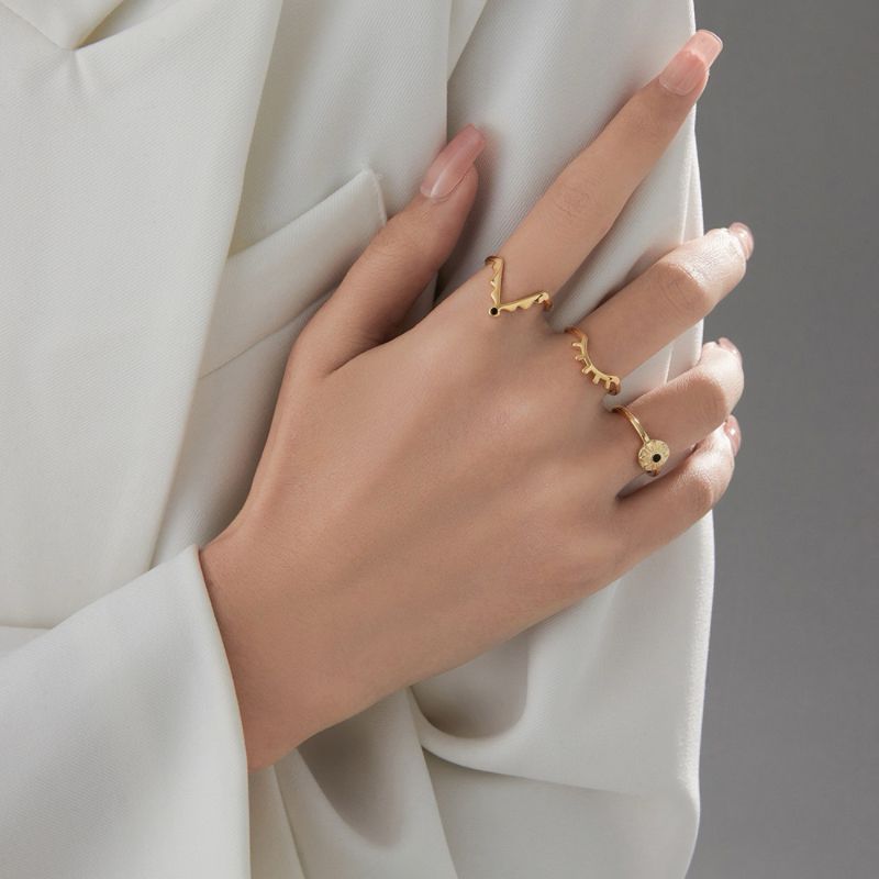Main de femme portant trois bagues plaquées or : une bague en V ornée d'une pierre noire, une bague torsadée et une bague œil bleu sertie de petites pierres.
