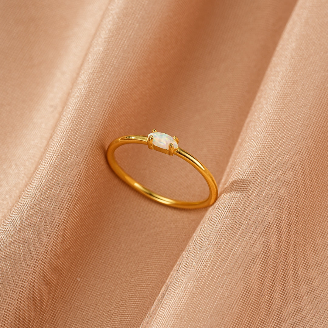 Bague fine en argent plaqué or avec une petite opale ovale au centre, posée sur un tissu satiné couleur pêche.