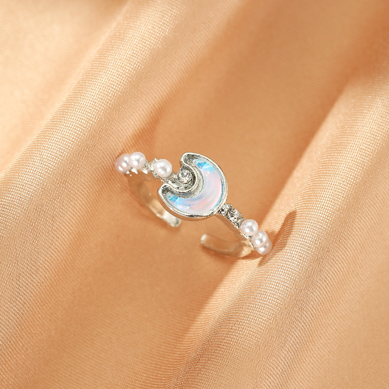 Bague ajustable argentée ornée d'une lune nacrée irisée, de petites perles blanches et de cristaux scintillants, posée sur un tissu satiné doré.